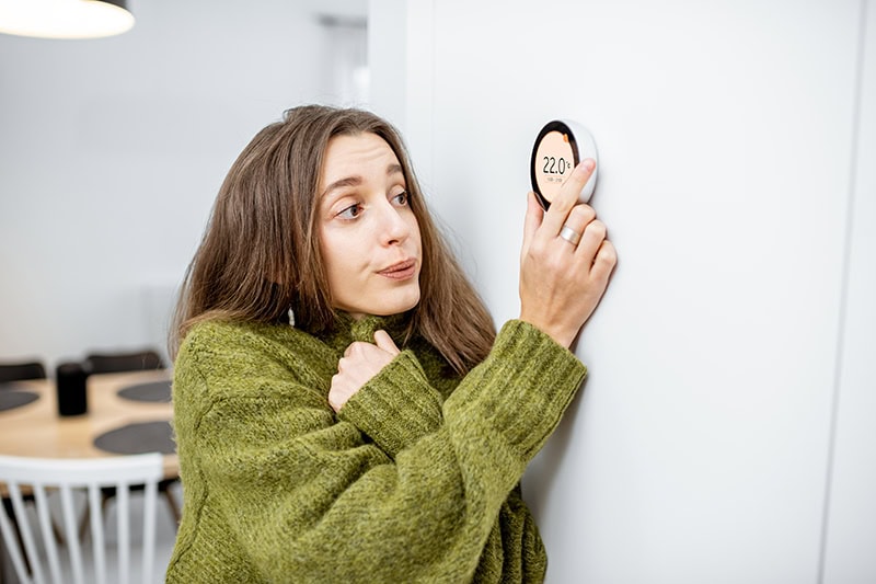 woman shivering in her sweater as she adjusts the thermostat in her home.